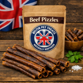 A kraft paper stand-up pouch labeled 'Beef Pizzles' by Beltin Pet Treats stands on a rustic wooden surface, with several beef jerky sticks placed in front, a small bowl of treats to the right, a green herb leaf, and a Union Jack flag blurred in the background.