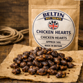 A stand-up pouch of Beltin Pet Treats Chicken Hearts snack stands on a rustic wooden surface with a pile of the dark brown coated treats placed in front on brown paper. The package label reads 'Beltin Pet Treats' and 'Chicken Hearts'.
