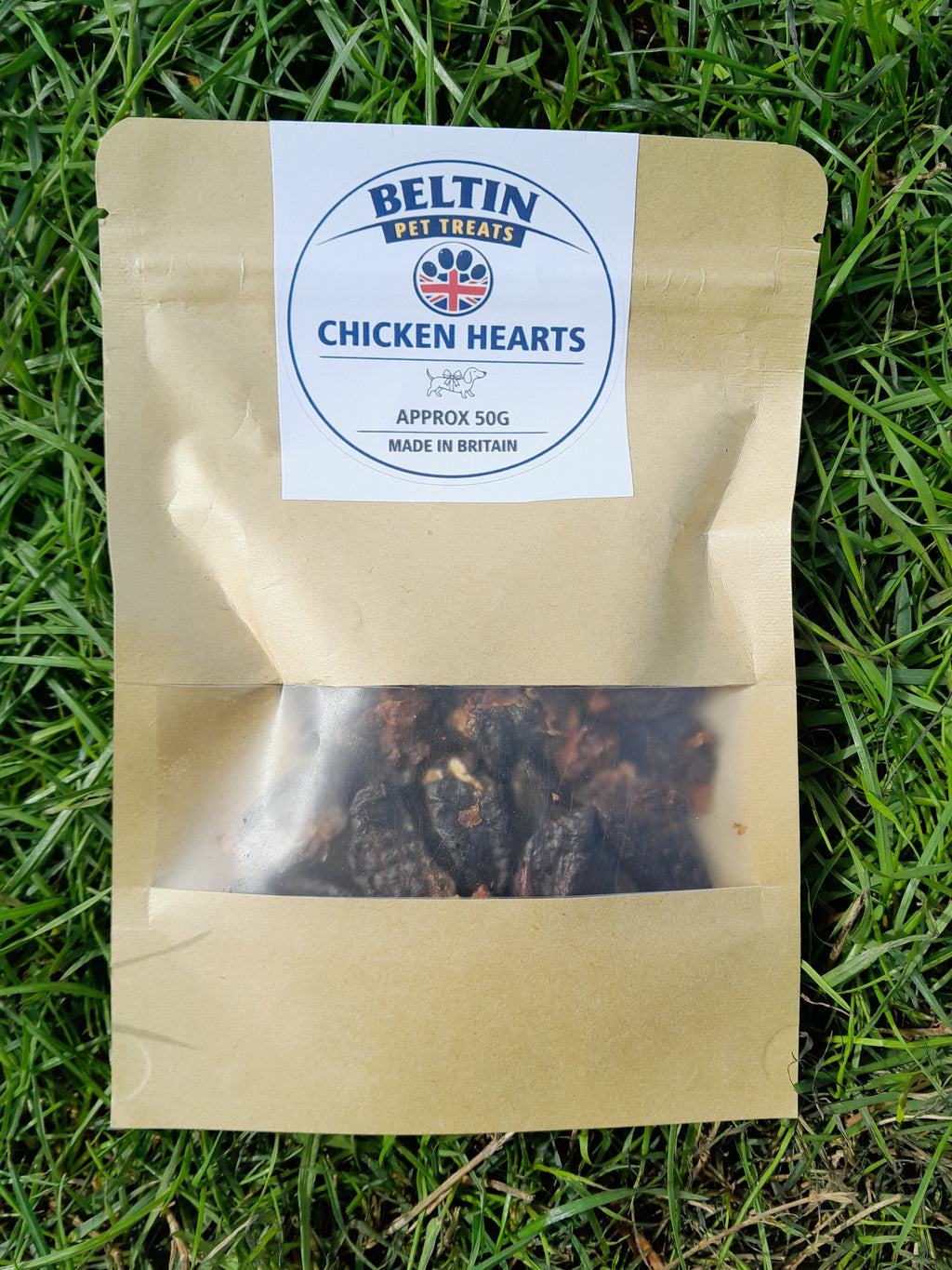 A kraft paper pouch of Bellin Chicken Hearts appetizers, placed on a green grass background. The pouch has a clear window showing the dark, textured appetizers inside, and a white circular label with the Bellin brand logo.