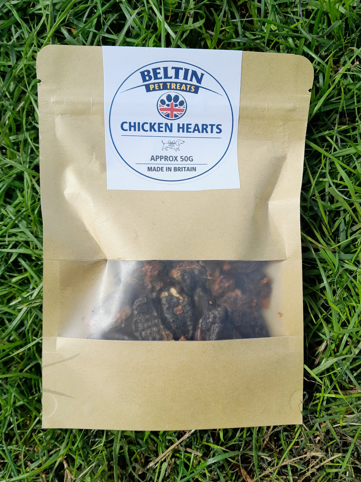 A kraft paper pouch of Bellin Chicken Hearts appetizers, placed on a green grass background. The pouch has a clear window showing the dark, textured appetizers inside, and a white circular label with the Bellin brand logo.