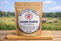 A stand-up pouch of Beltin Pet Treats Lamb Puffs 100% Lamb Lung treats, displayed on a wooden cutting board with a blurred outdoor field and sheep in the background.
