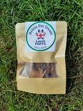 A bag of Belting Pups dog treats placed on green grass. The treat pouch is made of brown kraft paper with a clear window showing the treats inside, and a white circular label on the front with the brand name 'Belting Pups' and a Union Jack logo.
