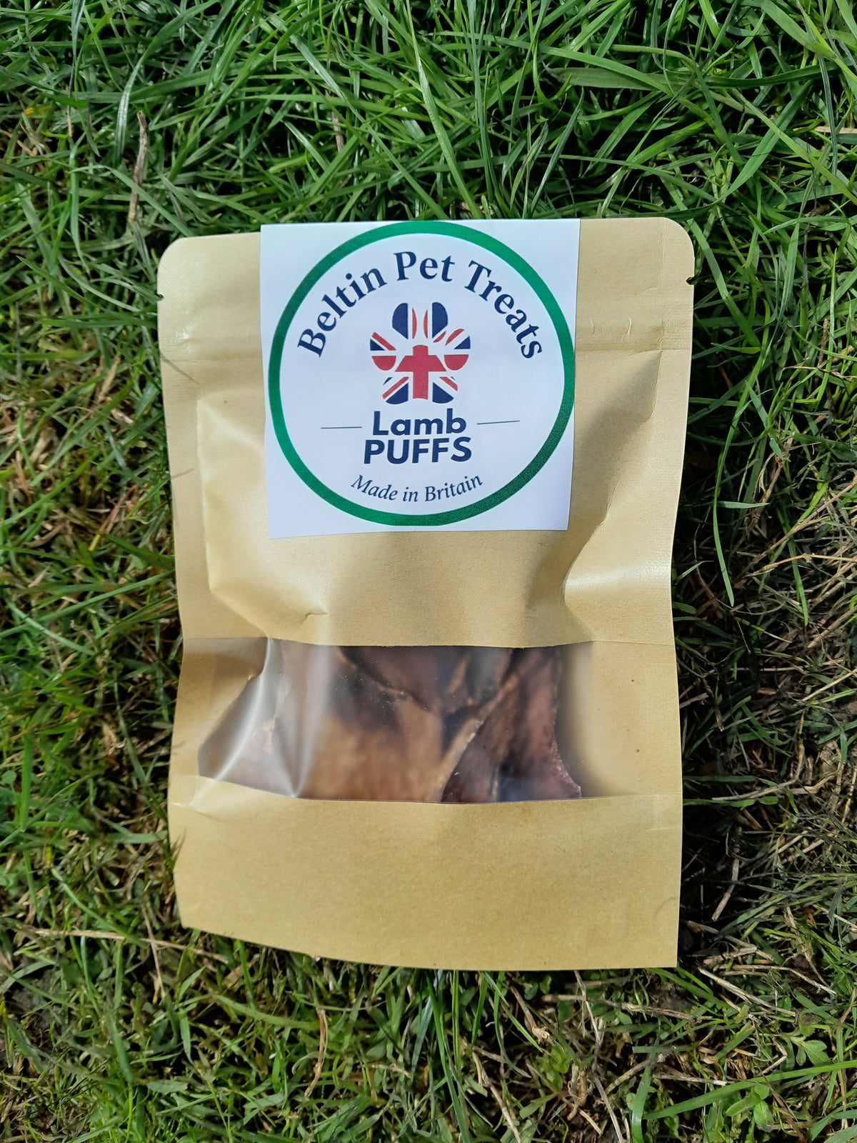 A bag of Belting Pups dog treats placed on green grass. The treat pouch is made of brown kraft paper with a clear window showing the treats inside, and a white circular label on the front with the brand name 'Belting Pups' and a Union Jack logo.